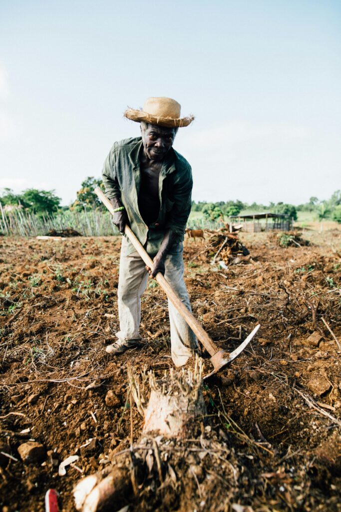 An African farmer wearing a straw hat tills the soil with a spade in a rural countryside field.